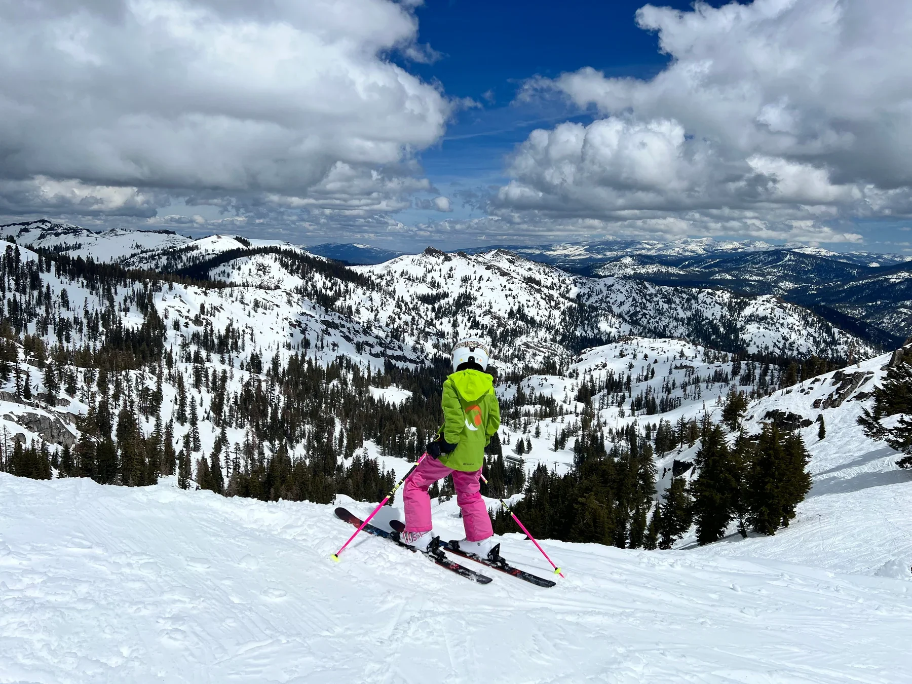 A skier in a bright green jacket and pink ski pants looking across wide snow-covered mountain slopes